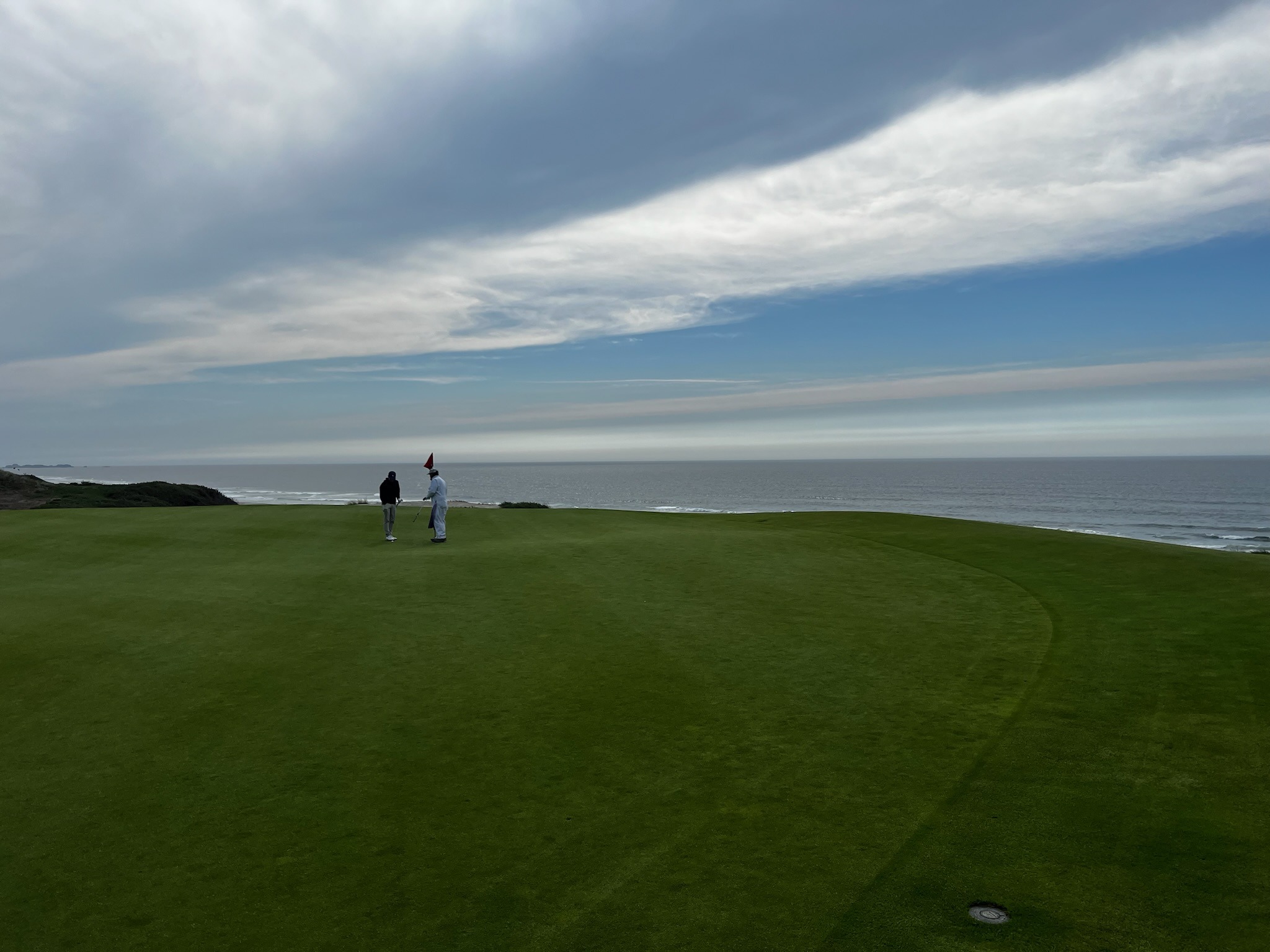 A caddie reading the line at the flag with a player on an oceanside green, guiding the putt to a clean hole-out
